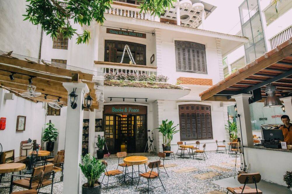 Khám Phá 9 Quán Cafe Độc Đáo Trên Đường Hoàng Hoa Thám 5 Quán cà phê ngoài trời với bàn ghế gỗ, cây xanh và biển hiệu "Books & Peace", không gian thư giãn và yên tĩnh.