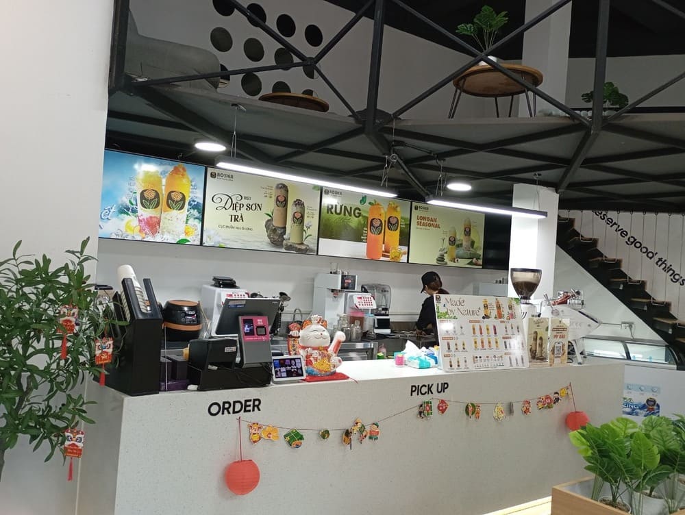 Khám Phá 15 Quán Cafe Hấp Dẫn Trên Đường Nguyễn Thị Định 13 Quầy phục vụ đồ uống với biển hiệu trang trí, nhân viên chuẩn bị đồ, cây xanh và cầu thang phía sau.
