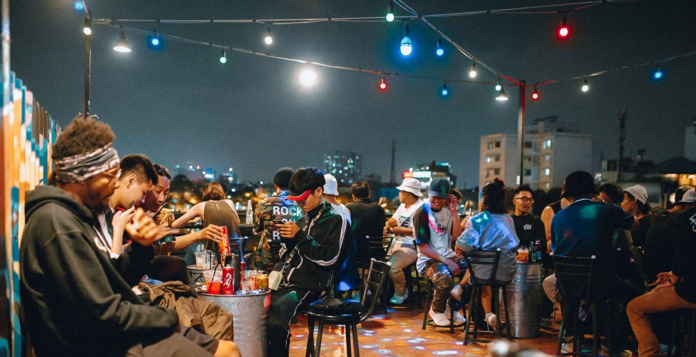 Khám Phá Top 10 Quán Rooftop Quận 1: View Đẹp Ngắm Thành Phố, Trải Nghiệm Thư Giãn Tuyệt Đỉnh 3 Nhóm người ngồi tại quán cà phê ngoài trời vào buổi tối, với đèn màu treo phía trên.