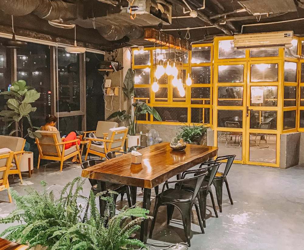 Khám Phá 7 Quán Cafe Tuyệt Đẹp Ở Ngã Tư Sở: View Đẹp, Đồ Uống Ngon, Giá Phải Chăng 15 Quán cà phê với bàn ghế gỗ, cây xanh trang trí và ánh sáng ấm áp, có người ngồi thư giãn gần cửa sổ.