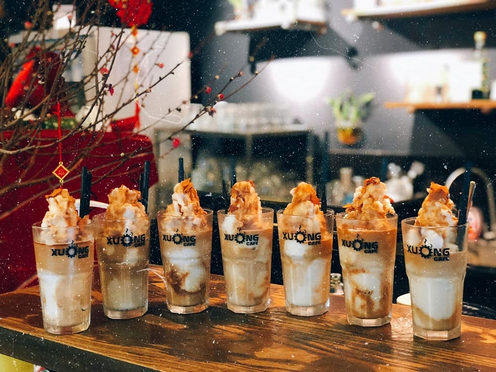 Khám Phá 15+ Quán Cafe View Đẹp, Đồ Uống Ngon Tại Hoàng Cầu 3 Bảy ly cà phê kem bày trên quầy gỗ, phía sau là không gian quán với cây trang trí.