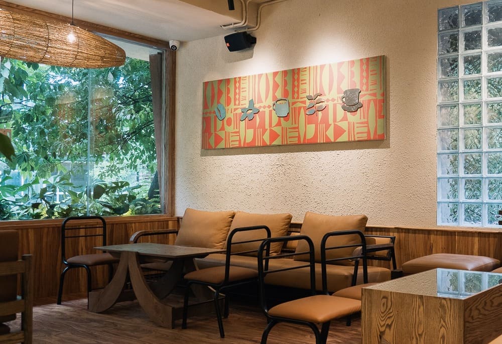 Khám Phá 15 Quán Cafe Hấp Dẫn Trên Đường Nguyễn Thị Định 12 Quán cà phê với ghế sofa màu nâu, bàn gỗ, tranh tường và cửa sổ nhìn ra cây xanh bên ngoài.