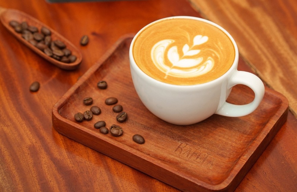 Khám Phá Top 10 Quán Cafe Giảng Võ Hấp Dẫn Nhất Tại Hà Nội 7 Tách cà phê latte với họa tiết nghệ thuật trên bề mặt, đặt trên khay gỗ, bên cạnh là hạt cà phê.