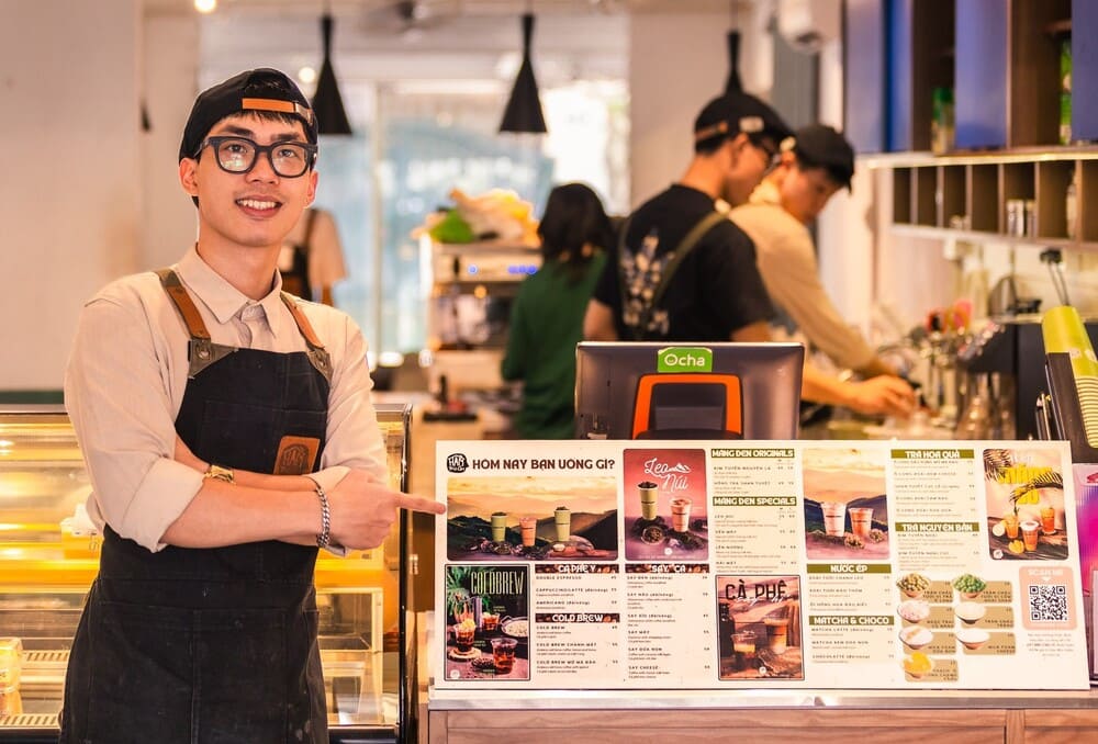 Khám Phá Top 10+ Quán Cafe Quán Thánh Lý Tưởng cho Hẹn Hò, Làm Việc và Thư Giãn 25 Nhân viên quán cà phê đứng bên quầy, chỉ vào thực đơn đồ uống với nhiều lựa chọn khác nhau.