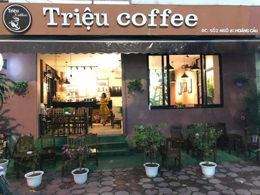 Khám Phá 15+ Quán Cafe View Đẹp, Đồ Uống Ngon Tại Hoàng Cầu 16 Quán cà phê với bảng hiệu "Triệu Coffee", có không gian bên trong ấm cúng và bài trí nhiều chậu cây xanh.