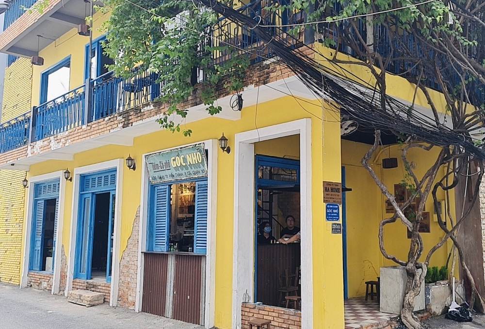 Khám Phá Những Quán Cafe Độc Lạ Thu Hút Giới Trẻ Tại Sài Gòn 9 Tòa nhà màu vàng với cửa xanh, dây leo phủ trên ban công, biển hiệu bên ngoài và người ngồi bên trong.