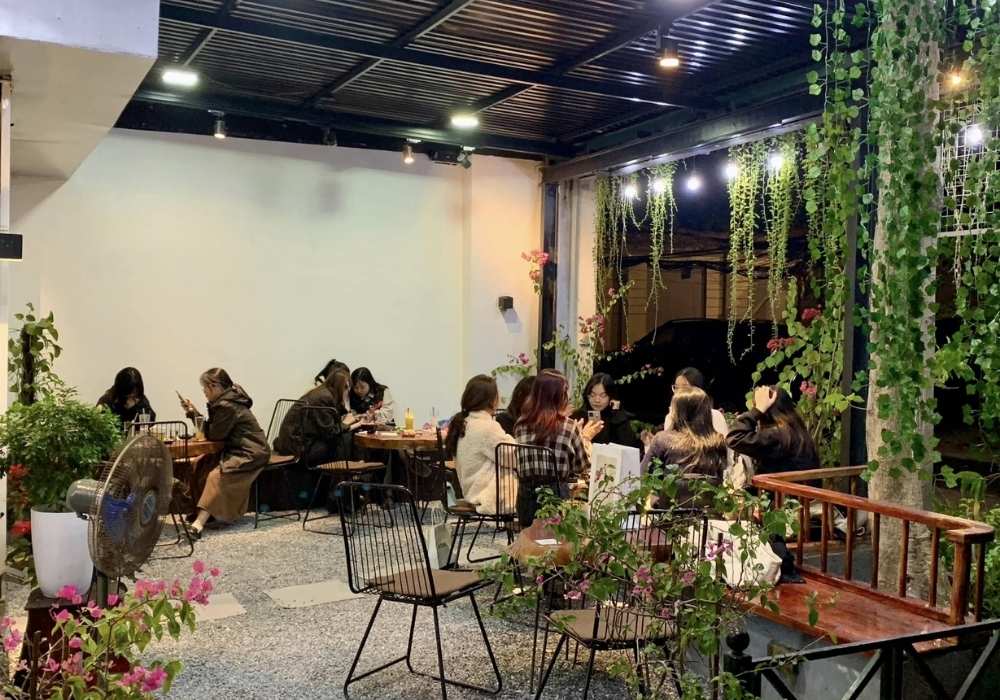 Khám Phá Top 10 Quán Cafe Láng Hạ: View Đẹp, Menu Hấp Dẫn, Giá Cực Hợp Lý 15 Quán cà phê ngoài trời với nhiều người ngồi trò chuyện, trang trí cây xanh và ánh sáng ấm áp.