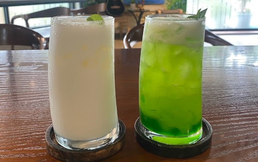 7 Quán Cafe View Đẹp Phạm Văn Đồng Không Thể Bỏ Qua 7 Hai ly nước uống, một màu trắng và một màu xanh, đặt trên bàn gỗ trong quán cà phê.