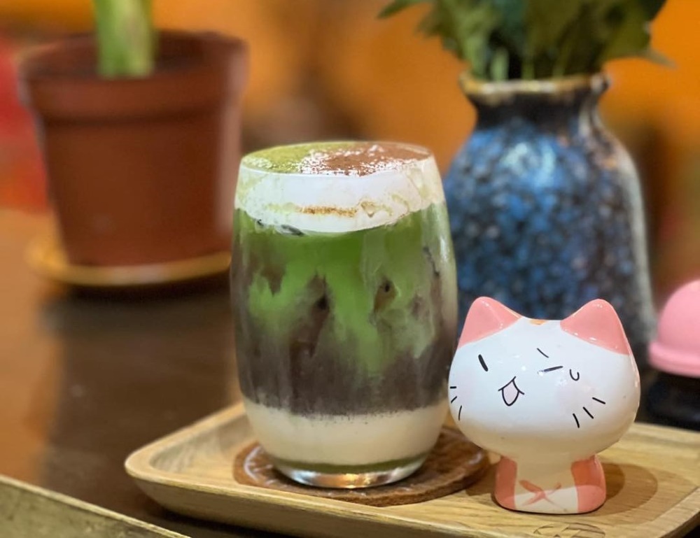 Khám Phá Top 10 Quán Cafe Giảng Võ Hấp Dẫn Nhất Tại Hà Nội 12 Ly đồ uống ba lớp màu xanh, nâu, trắng trên khay gỗ cạnh mô hình mèo nhỏ.