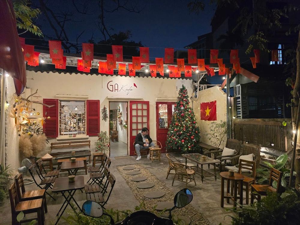 Khám Phá 10+ Quán Cafe Đẹp Ở Đặng Văn Ngữ: Đồ Uống Ngon, Giá Cả Hấp Dẫn 9 Quán cà phê trang trí cờ và cây thông, có người ngồi bên ngoài vào buổi tối.