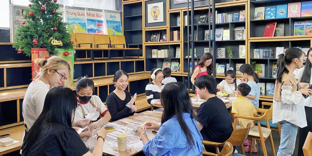 Khám Phá Top Quán Cafe Sách Hot Nhất Quận 1 Bạn Không Thể Bỏ Lỡ Năm Nay 11 Nhóm người đang tham gia hoạt động thủ công trong một thư viện có trang trí cây thông Noel và giá sách.