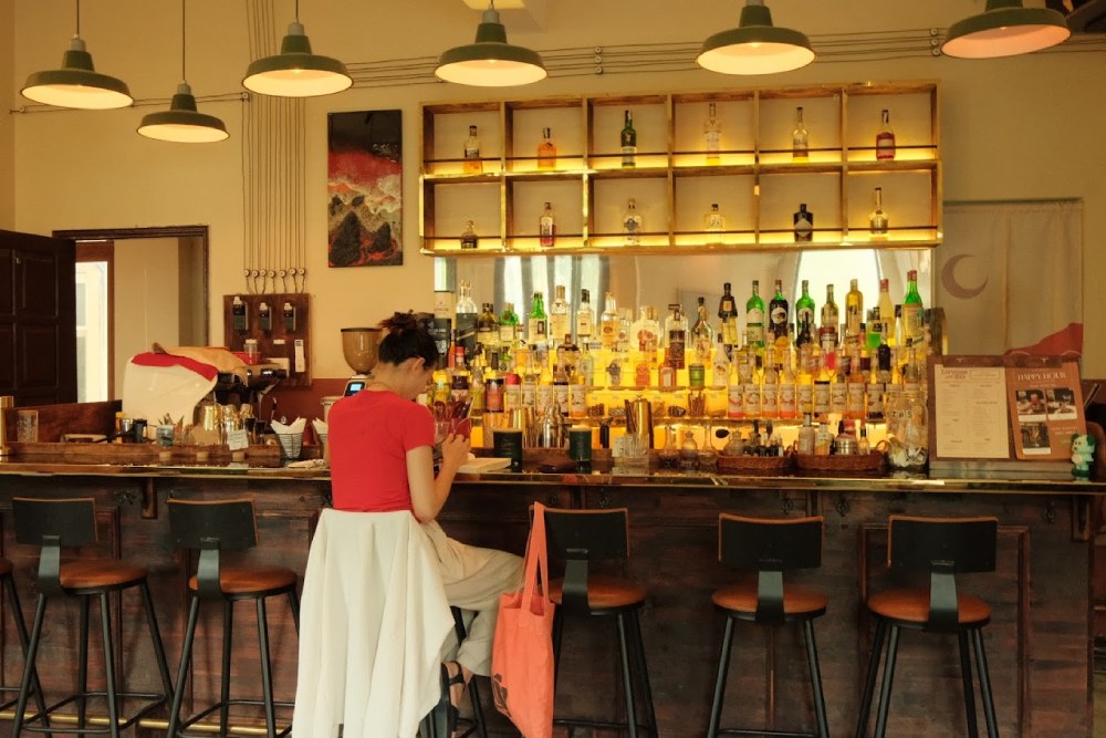 Khám Phá 11 Quán Cafe Đội Cấn Tuyệt Vời Cho Thư Giãn Và Làm Việc 16 Người ngồi tại quầy bar với nhiều chai rượu trên kệ, ánh sáng ấm áp trong không gian quán.