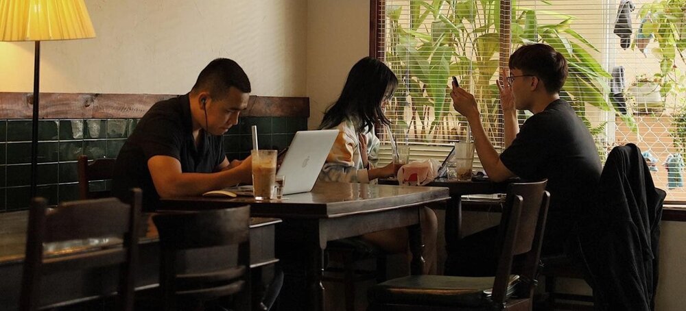 Khám Phá 9 Quán Cafe Độc Đáo Trên Đường Hoàng Hoa Thám 21 Ba người ngồi trong quán cà phê, mỗi người sử dụng thiết bị điện tử riêng, ánh sáng từ cửa sổ chiếu vào.