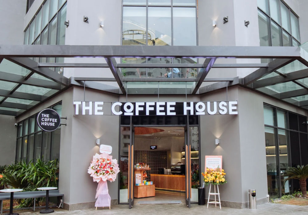 Khám Phá Top 6 Quán Cafe Hot Nhất Tại Thành Thái, Cầu Giấy 12 Mặt tiền quán cà phê với bảng hiệu lớn, cửa kính và hai kệ hoa tươi bên ngoài, nằm trong tòa nhà hiện đại.