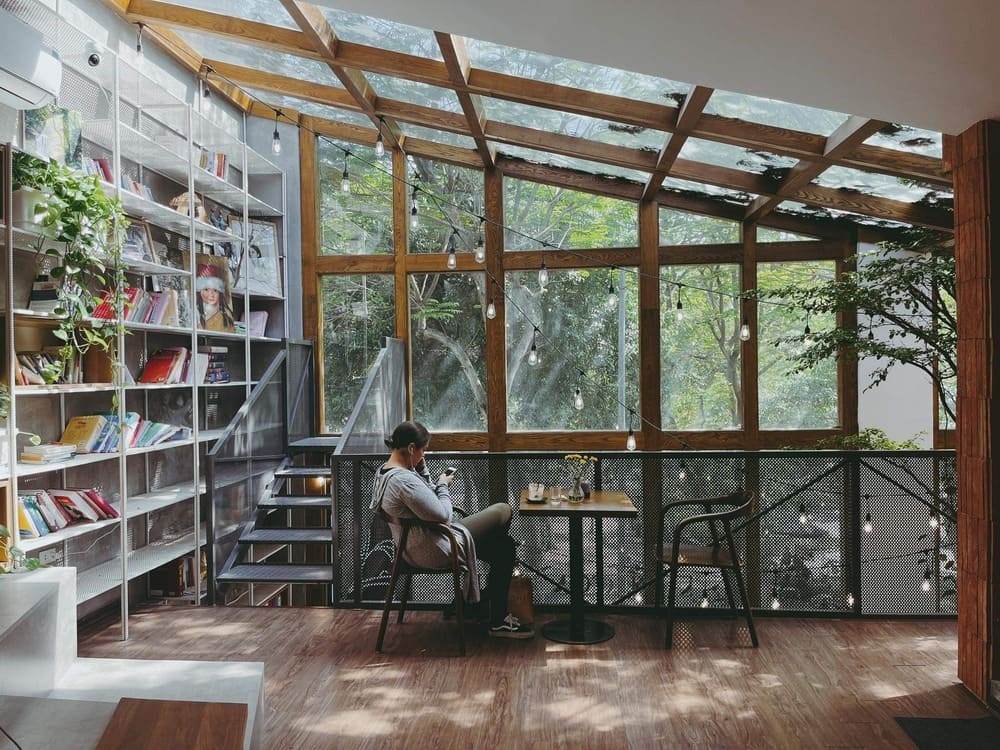 Khám Phá Top 10+ Quán Cafe Quán Thánh Lý Tưởng cho Hẹn Hò, Làm Việc và Thư Giãn 5 Người ngồi trong quán cà phê có cửa sổ lớn, ánh sáng tự nhiên chiếu vào, bên cạnh là kệ sách và cây xanh.