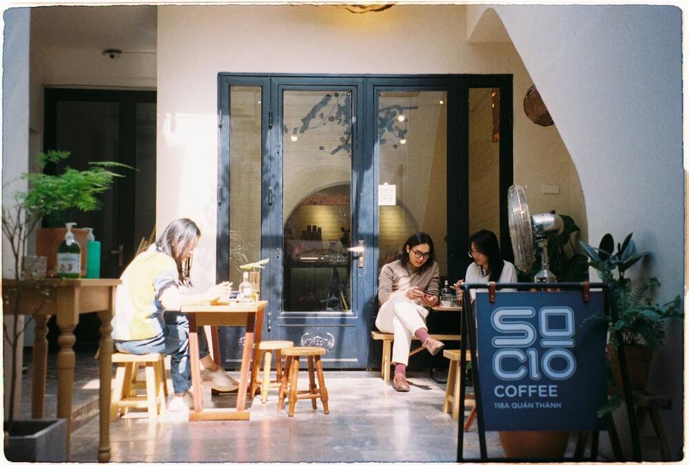 Khám Phá Top 10+ Quán Cafe Quán Thánh Lý Tưởng cho Hẹn Hò, Làm Việc và Thư Giãn 11 Ba người ngồi bên ngoài quán cà phê, một người đọc sách, hai người khác sử dụng điện thoại, cạnh biển hiệu quán.