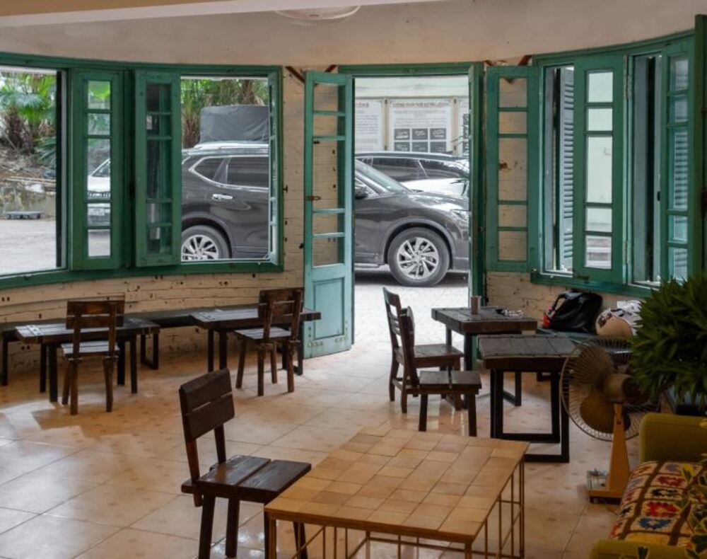 Khám Phá 9 Quán Cafe Độc Đáo Trên Đường Hoàng Hoa Thám 14 Quán cà phê với bàn ghế gỗ, cửa sổ mở ra ngoài, hai chiếc xe đỗ bên ngoài, không gian thoáng đãng.