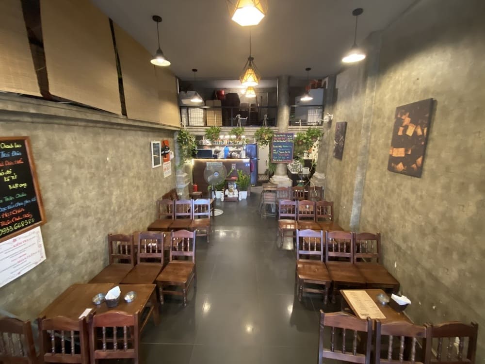 Khám Phá Top 10+ Quán Cafe Quán Thánh Lý Tưởng cho Hẹn Hò, Làm Việc và Thư Giãn 19 Quán cà phê có bàn ghế gỗ, trang trí cây xanh, ánh sáng đèn vàng, không gian ấm cúng và thực đơn trên tường.