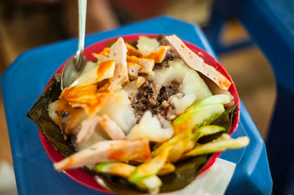 Món bánh giò trên đĩa đỏ, trang trí với giò lụa và dưa leo, kèm theo muỗng, đặt trên bàn màu xanh.