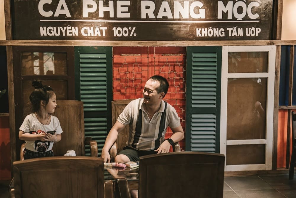 Khám Phá 15 Quán Cafe Hấp Dẫn Trên Đường Nguyễn Thị Định 10 Người đàn ông và bé gái ngồi trò chuyện vui vẻ trong quán cà phê với biển hiệu "Cà Phê Rang Mộc".
