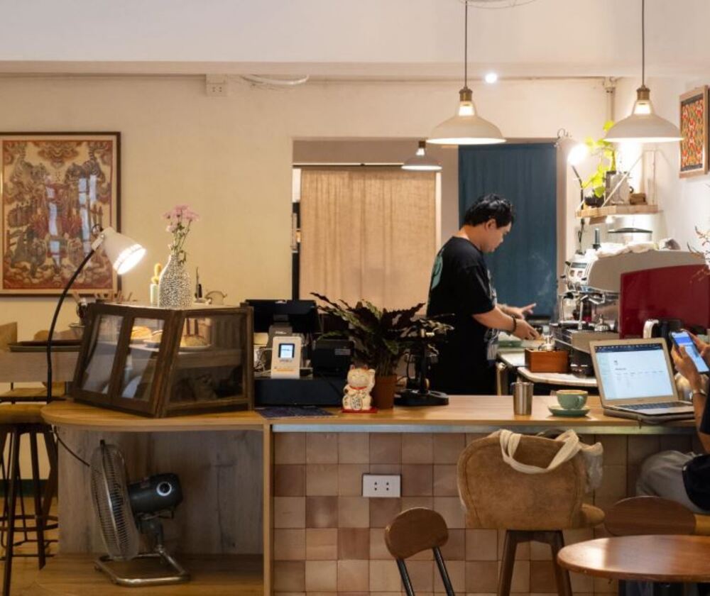 Khám Phá 9 Quán Cafe Độc Đáo Trên Đường Hoàng Hoa Thám 13 Quán cà phê ấm cúng với nhân viên pha chế, máy pha cà phê, bàn làm việc có laptop và trang trí nhẹ nhàng.