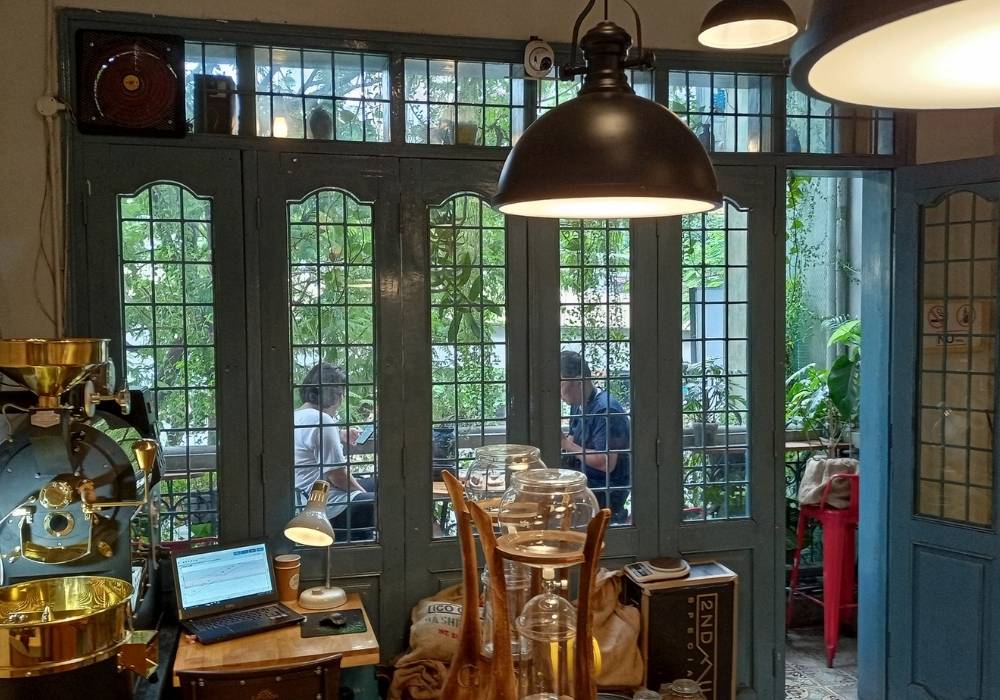 Khám Phá Top 10 Quán Cafe Láng Hạ: View Đẹp, Menu Hấp Dẫn, Giá Cực Hợp Lý 8 Quán cà phê có cửa sổ lớn, ánh sáng ấm áp, máy tính trên bàn, người ngồi ngoài ban công nhìn vào.