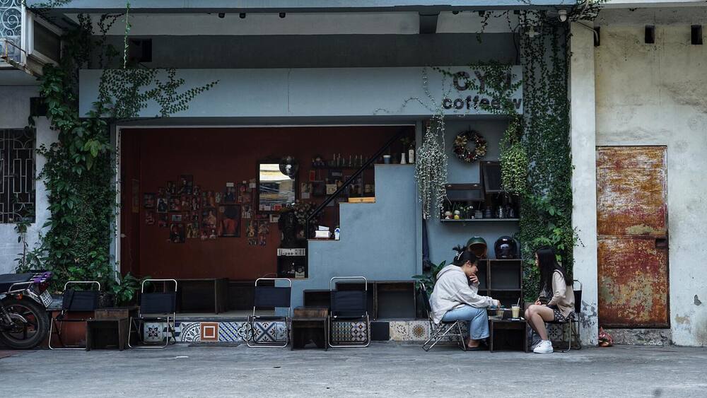 Khám Phá 9 Quán Cafe Độc Đáo Trên Đường Hoàng Hoa Thám 7 Hai người ngồi uống cà phê trước quán, với cây xanh và đồ trang trí treo tường xung quanh.