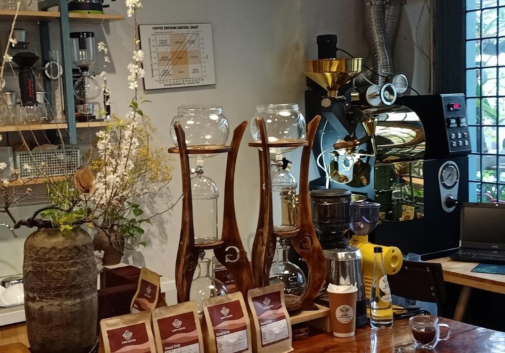 Khám Phá Top 10 Quán Cafe Láng Hạ: View Đẹp, Menu Hấp Dẫn, Giá Cực Hợp Lý 7 Nội thất quán cà phê với máy rang xay, bình hoa, và các sản phẩm cà phê trên bàn gỗ.