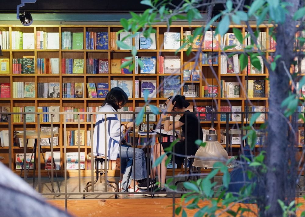 Khám Phá 11 Quán Cafe Sách Độc Đáo Tại Cầu Giấy Hút Hồn Độc Giả 18 Hai người ngồi đọc sách trong một quán cà phê sách, xung quanh là kệ sách đầy màu sắc.