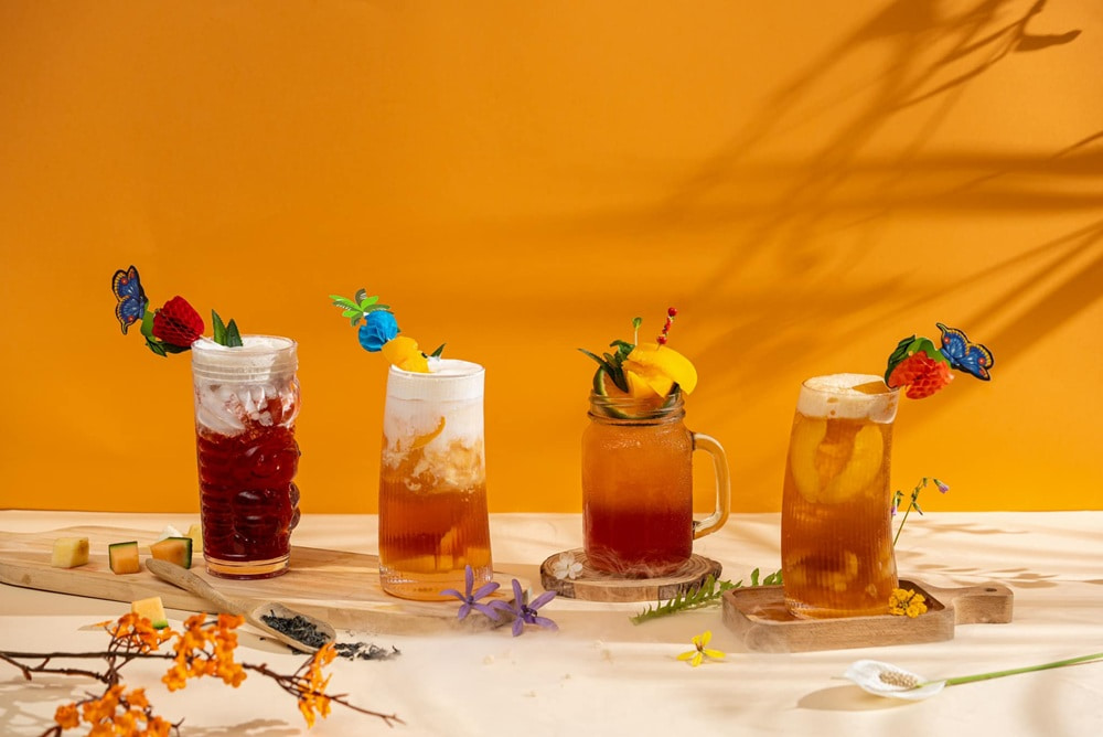 Khám Phá 15+ Quán Cafe View Đẹp, Đồ Uống Ngon Tại Hoàng Cầu 8 Bốn ly nước giải khát trang trí với trái cây và hình bướm, nền màu cam sáng, ánh sáng tự nhiên.