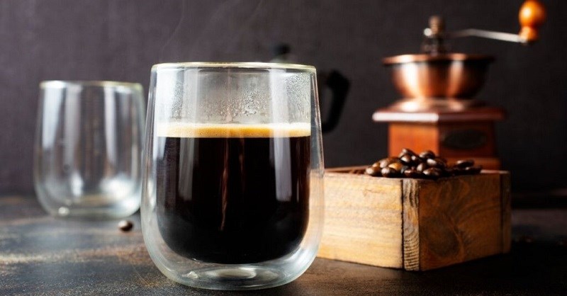 ca-phe-americano-la-gi Ly cà phê đen trên bàn, bên cạnh là hạt cà phê và máy xay cổ điển, tạo cảm giác ấm cúng và thư giãn.