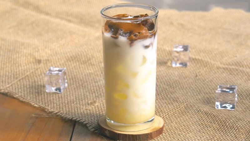 Ly cà phê sữa đá trên nền vải, kèm vài viên đá nhỏ xung quanh, tạo cảm giác mát mẻ và hấp dẫn.