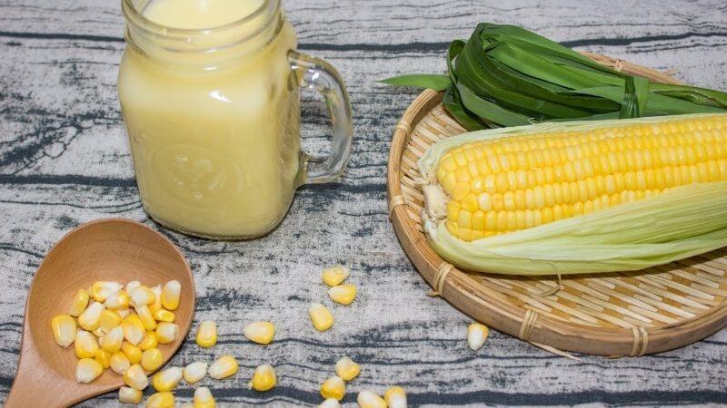 Ngô trên rổ, ly sữa ngô, và hạt ngô trên thìa gỗ, đặt trên bề mặt bàn màu xám.