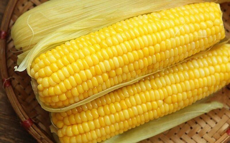 Hai bắp ngô vàng tươi nằm trên rổ tre, với lớp vỏ xanh nhạt ở phía dưới.