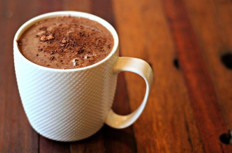 Tách sô cô la nóng trong cốc sứ trắng trên bàn gỗ, có lớp bột cacao rắc phía trên.