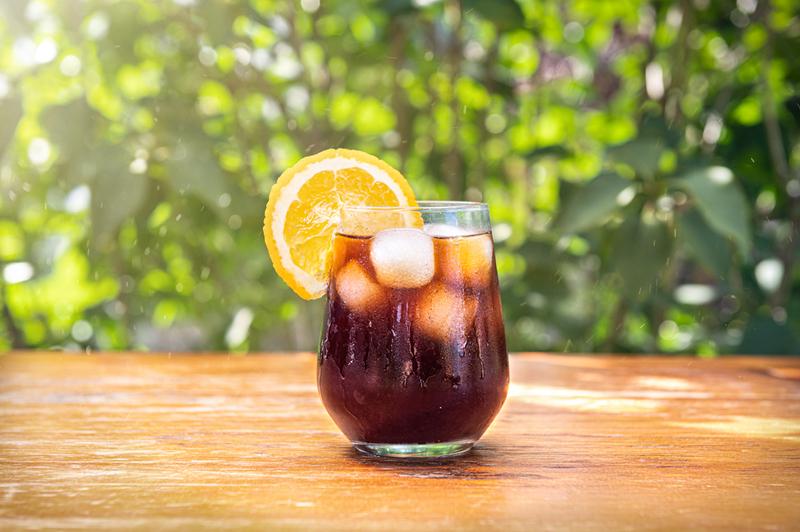 Ly nước lạnh với đ&aacute; v&agrave; l&aacute;t cam tr&ecirc;n b&agrave;n gỗ, nền l&agrave; c&acirc;y xanh.