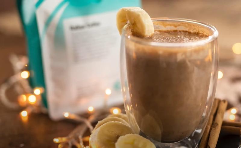 Ly sinh tố chuối trang tr&iacute; l&aacute;t chuối, đặt gần t&uacute;i bột v&agrave; đ&egrave;n trang tr&iacute; tr&ecirc;n b&agrave;n gỗ.