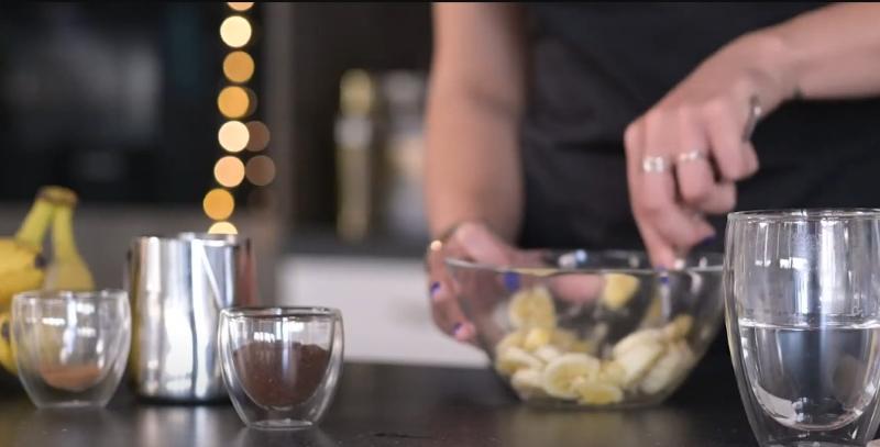 Người đang trộn chuối trong b&aacute;t thủy tinh, b&ecirc;n cạnh l&agrave; ly c&agrave; ph&ecirc;, b&igrave;nh pha v&agrave; tr&aacute;i chuối tr&ecirc;n b&agrave;n.