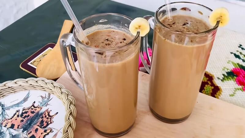 Hai ly c&agrave; ph&ecirc; sữa đ&aacute; với l&aacute;t chuối trang tr&iacute;, đặt tr&ecirc;n b&agrave;n c&ugrave;ng một chiếc đĩa v&agrave; dụng cụ bằng gỗ.