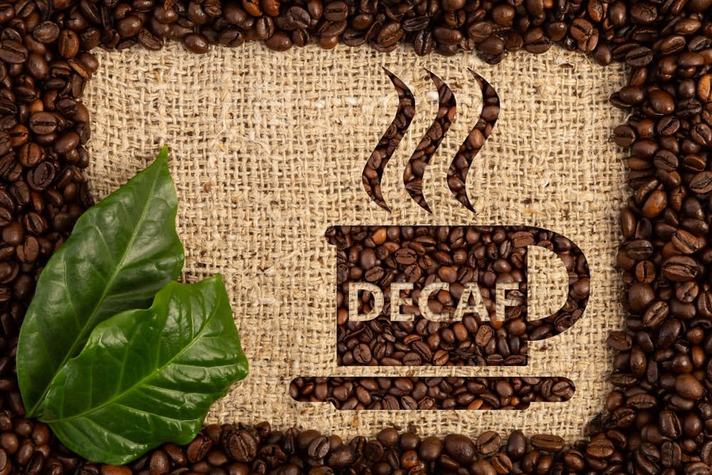 ca-phe-decaf-la-gi Hạt cà phê và lá xanh trên nền vải bố, hình tách cà phê chữ "DECAF" được tạo bằng hạt cà phê.