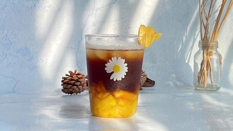 Ly nước lạnh với l&aacute;t dứa trang tr&iacute;, đặt tr&ecirc;n mặt b&agrave;n, c&oacute; nền s&aacute;ng v&agrave; một số đồ trang tr&iacute; tự nhi&ecirc;n xung quanh.