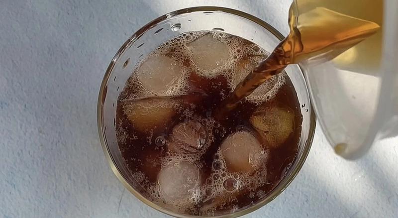 Ly nước đ&aacute; đang được r&oacute;t th&ecirc;m nước m&agrave;u n&acirc;u từ một b&igrave;nh, nh&igrave;n từ tr&ecirc;n xuống.