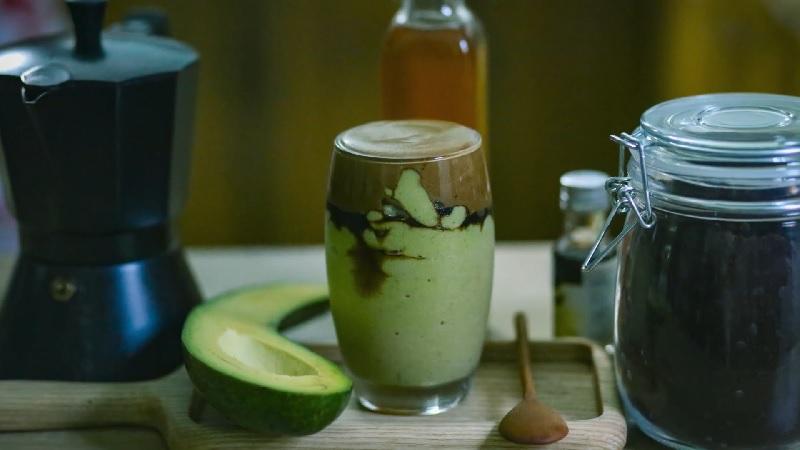 Ly sinh tố bơ với lớp socola v&agrave; c&agrave; ph&ecirc;, b&ecirc;n cạnh l&agrave; nửa quả bơ, b&igrave;nh c&agrave; ph&ecirc;, v&agrave; hũ thủy tinh tr&ecirc;n b&agrave;n.