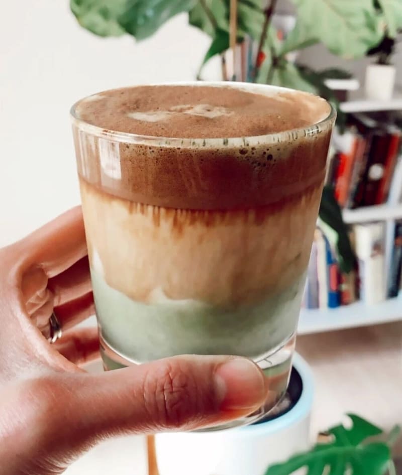 Người cầm ly c&agrave; ph&ecirc; đ&aacute; xay nhiều tầng, ph&iacute;a sau l&agrave; kệ s&aacute;ch v&agrave; c&acirc;y xanh.