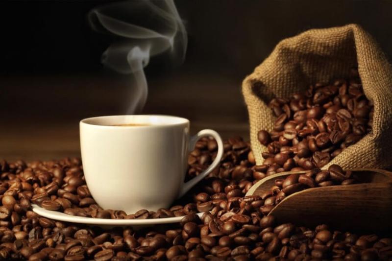 Tách cà phê nóng bốc khói đặt trên đĩa, xung quanh là hạt cà phê và túi vải mở đựng hạt.