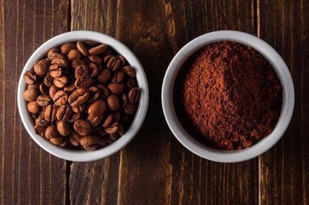 Hai chén trên bàn gỗ, một chứa hạt cà phê và một chứa cà phê xay nhuyễn.