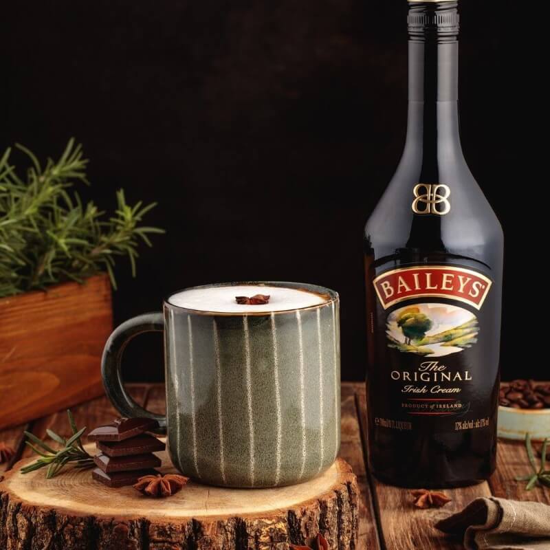 Cốc c&agrave; ph&ecirc; cạnh chai rượu Baileys tr&ecirc;n nền gỗ với c&acirc;y xanh v&agrave; thanh s&ocirc;-c&ocirc;-la trang tr&iacute; xung quanh.