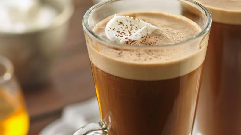 Ly c&agrave; ph&ecirc; n&oacute;ng với lớp bọt sữa v&agrave; kem tươi, trang tr&iacute; &iacute;t bột cacao, đặt tr&ecirc;n b&agrave;n gỗ.