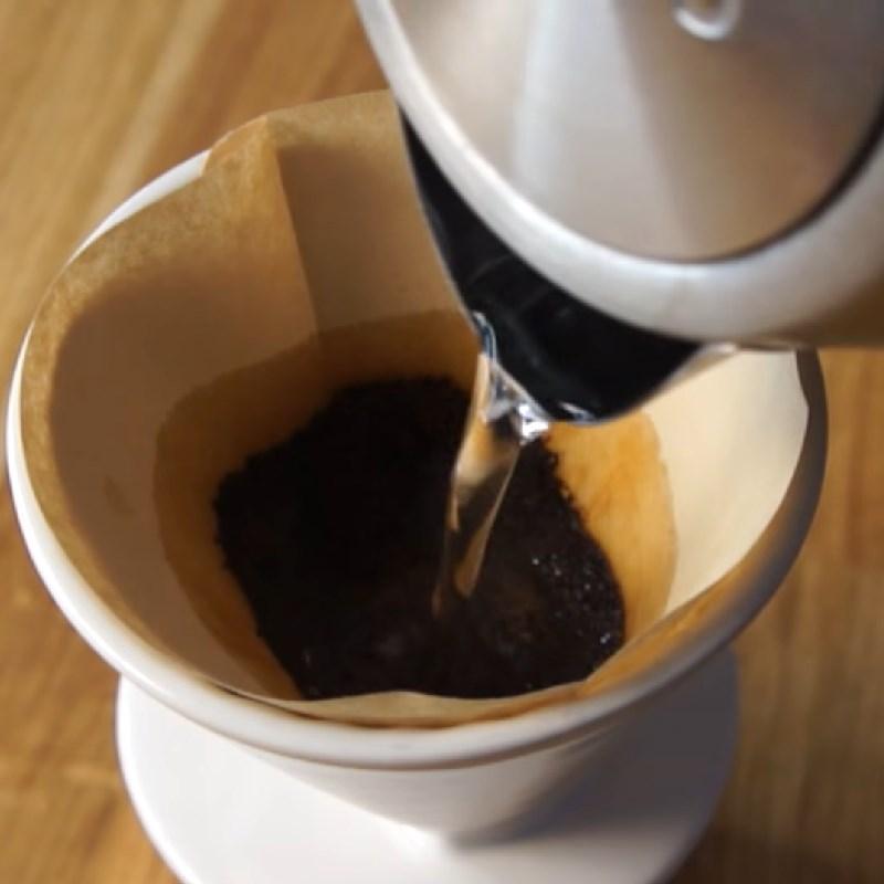 Nước đang được r&oacute;t v&agrave;o phin c&agrave; ph&ecirc; để pha chế, với bột c&agrave; ph&ecirc; đặt trong giấy lọc tr&ecirc;n nền gỗ.