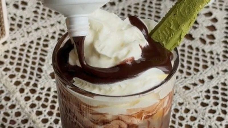 Ly kem phủ sốt socola v&agrave; kem tươi, trang tr&iacute; với một thanh b&aacute;nh vị tr&agrave; xanh, tr&ecirc;n nền khăn ren.